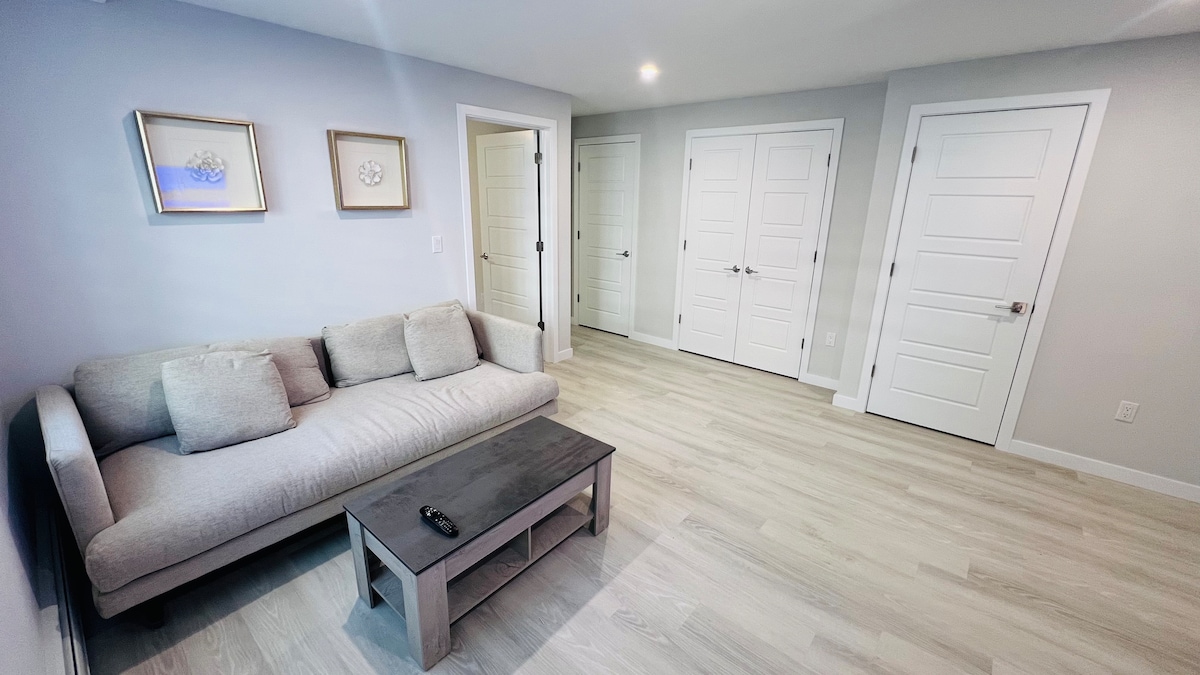 A light-colored living area features a comfortable sectional sofa with decorative pillows in front of a sleek coffee table. Two closed doors provide privacy, while additional doors lead to bedrooms. Soft, natural light enhances the neutral wall tones and the pale wood flooring.