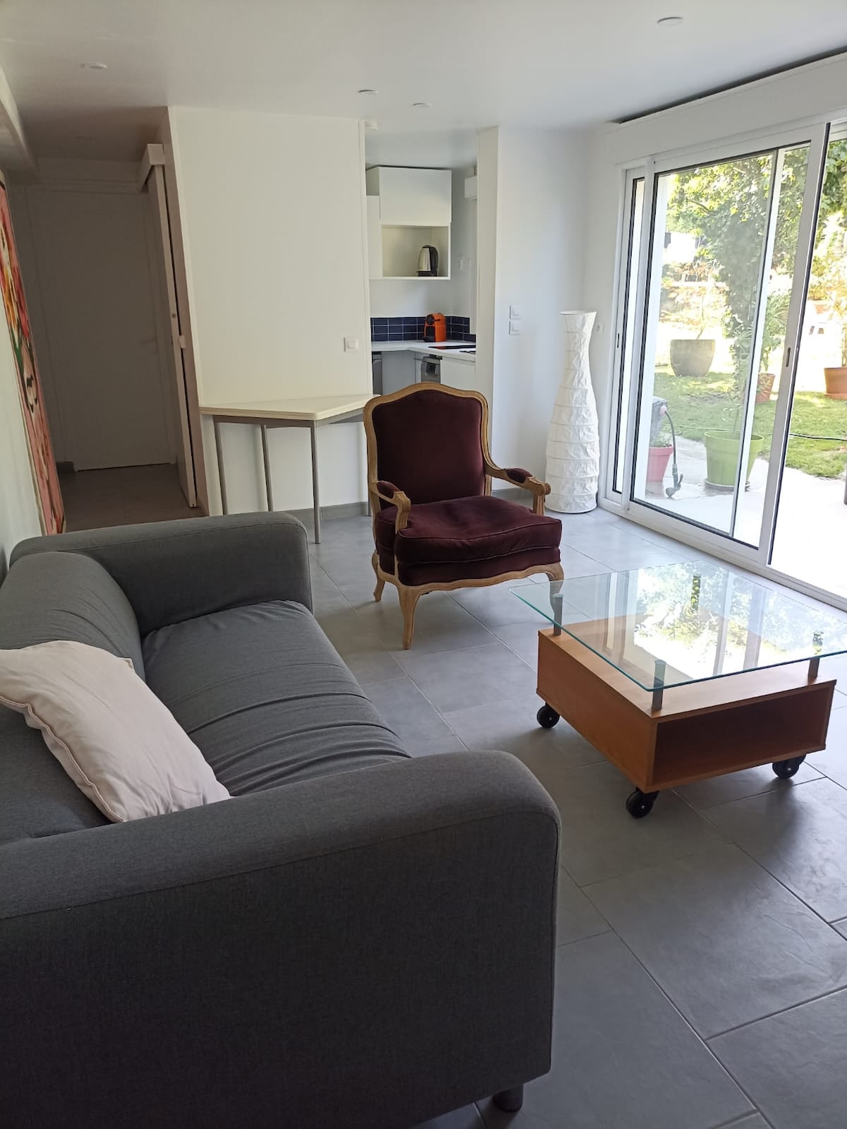 A spacious living area is present, featuring a gray sofa positioned beside a glass coffee table on wheels. A burgundy armchair adds a touch of elegance. Large glass doors provide natural light and a view into a shared garden space.