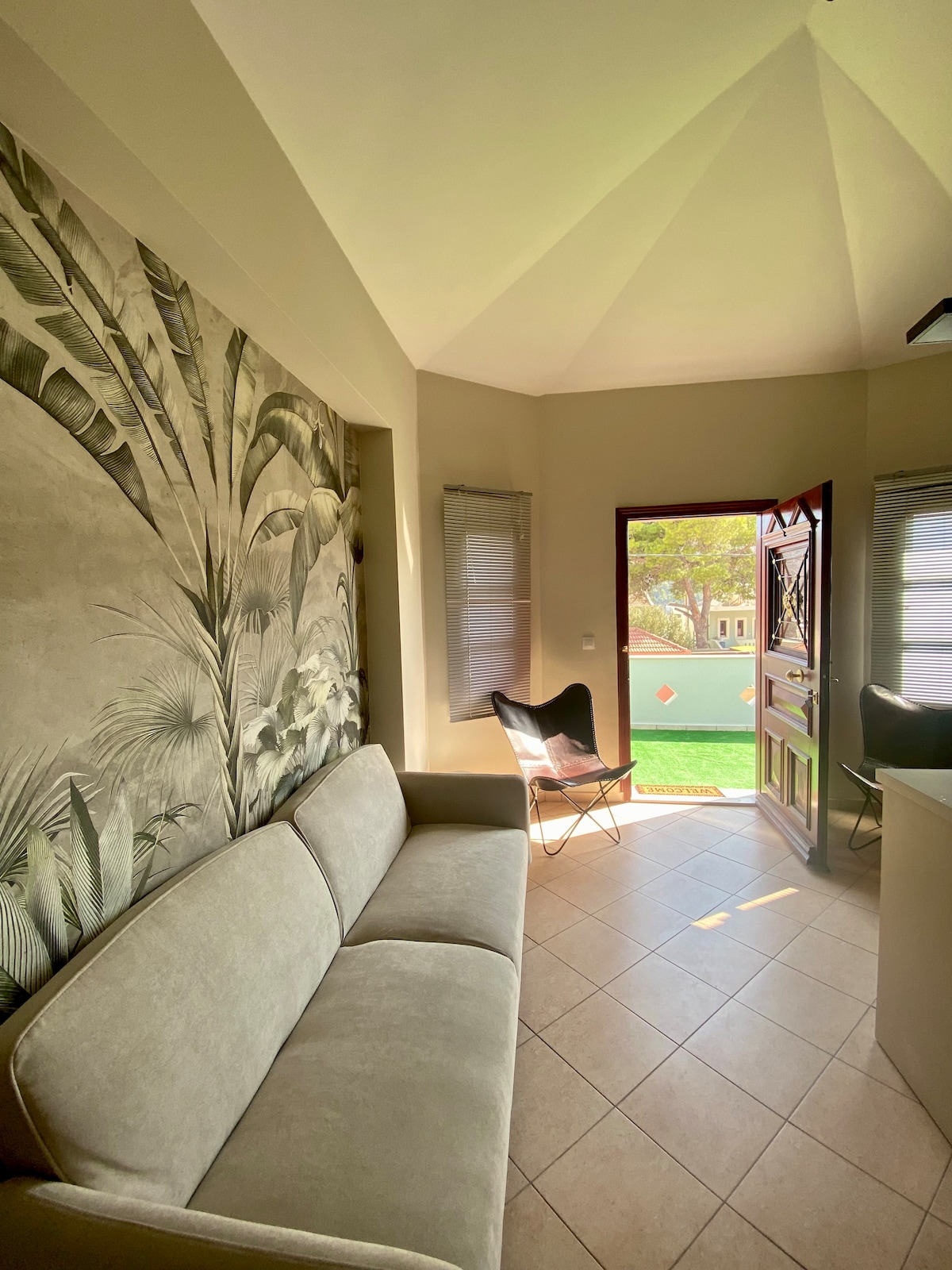 A light-filled living area features a neutral sofa against a botanical wall mural. An open door reveals a glimpse of the outdoor space, complemented by two softly shaded windows that enhance the serene atmosphere.