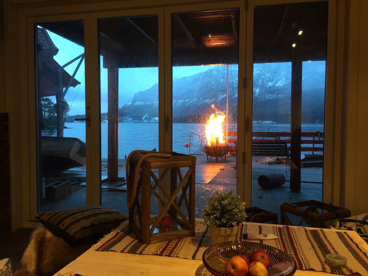Large glass doors provide a view of the tranquil water and a flickering fire pit on the deck. Soft lighting is visible through the window, reflecting a cozy indoor space with a wooden table and decorative elements like a plant and a bowl of apples.