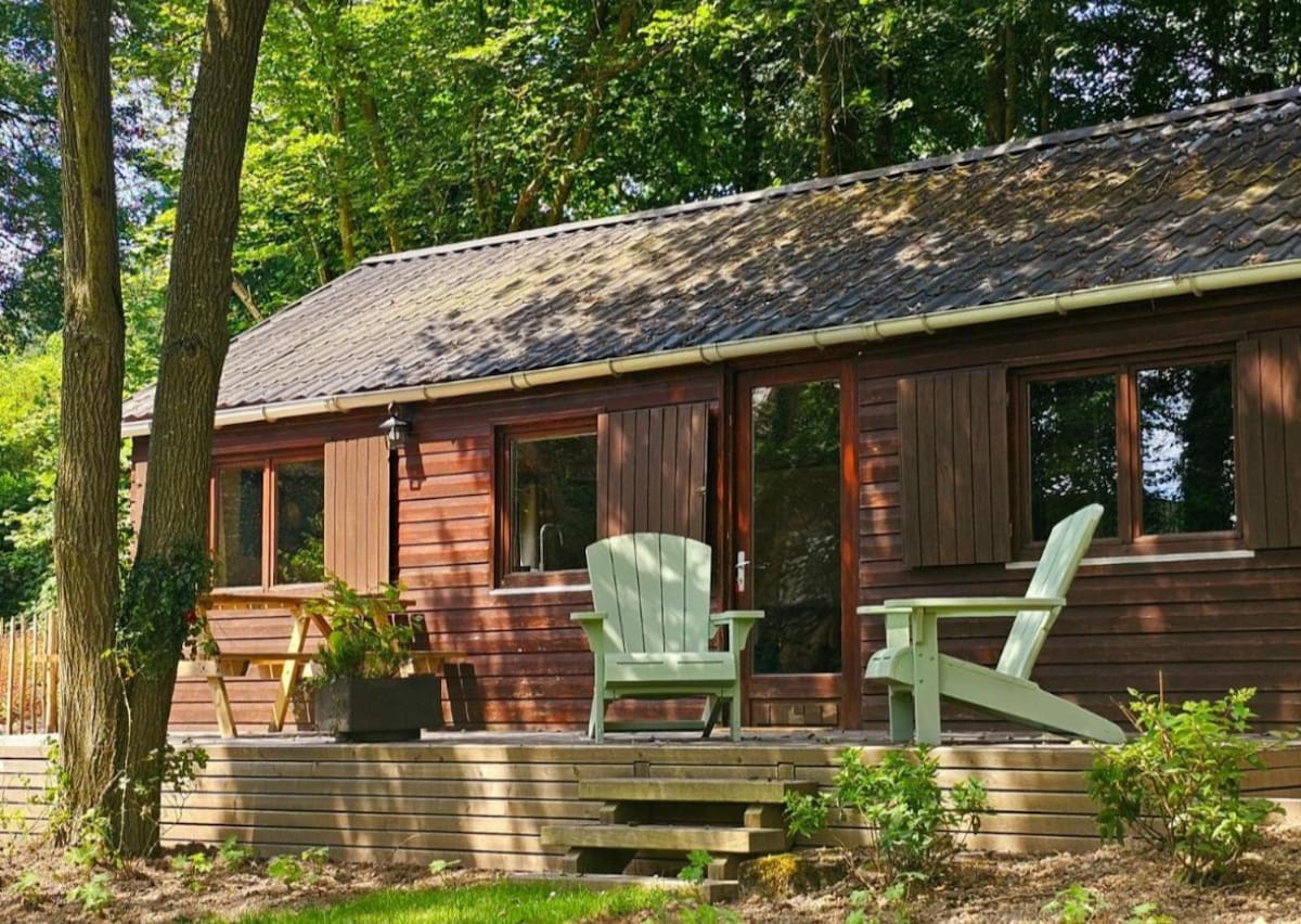 A charming wooden cabin is nestled among trees, featuring large windows framed by shutters. Two light green chairs are placed on the deck, complemented by a small table and potted plant, creating an inviting outdoor space for relaxation.