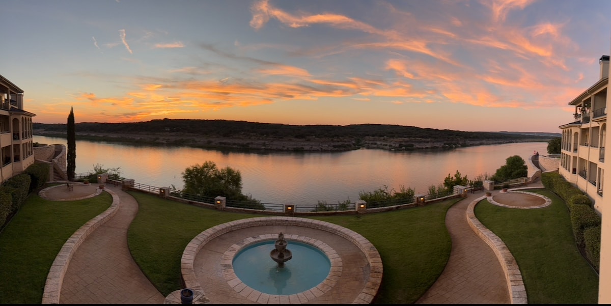 A panoramic view showcases a serene lake at sunset, reflecting shades of orange and purple in the water. Well-maintained green lawns frame a circular fountain in the foreground, while gentle hills and a row of trees outline the distant shore.