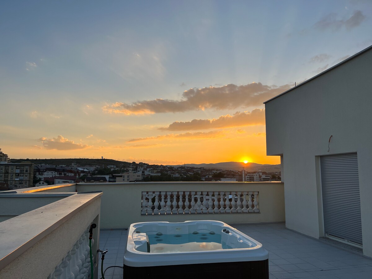 A rooftop terrace is featured, showcasing a hot tub surrounded by a tiled floor. The sunset casts warm hues across the sky, with distant hills and cityscape visible in the background. A railing adds an open feel to the space, enhancing the view.