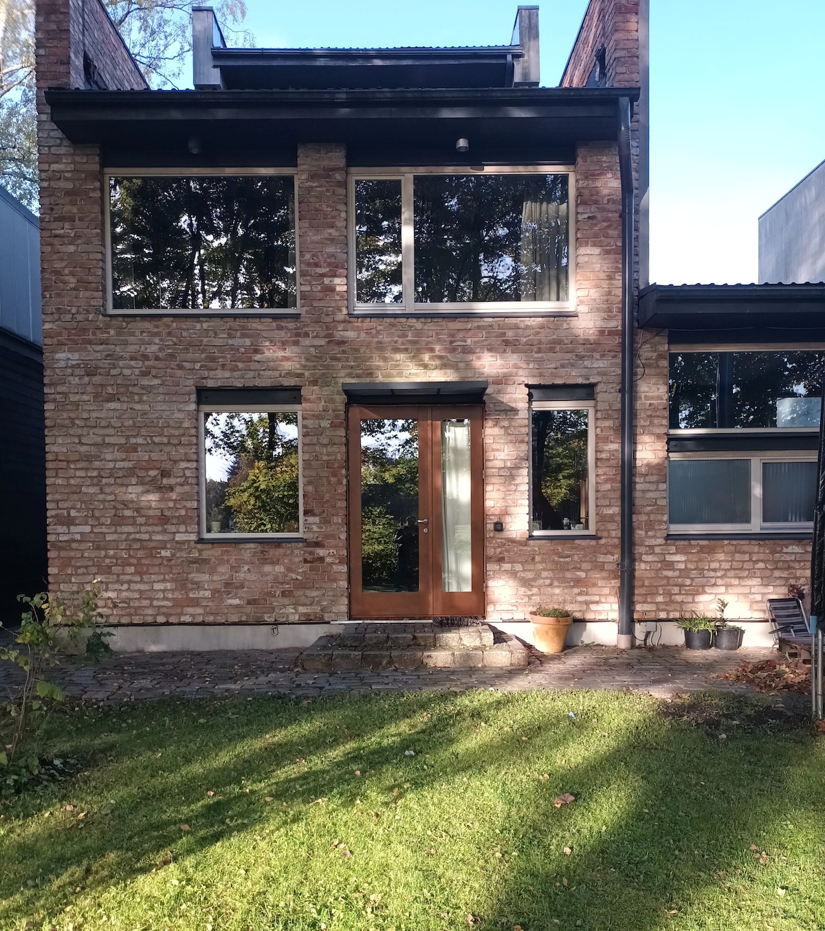 The exterior of the home showcases a combination of brick and modern materials, featuring large windows that allow natural light to enter. A wooden door serves as the entrance, framed by small planters. Lush greenery surrounds the building, enhancing the serene environment.
