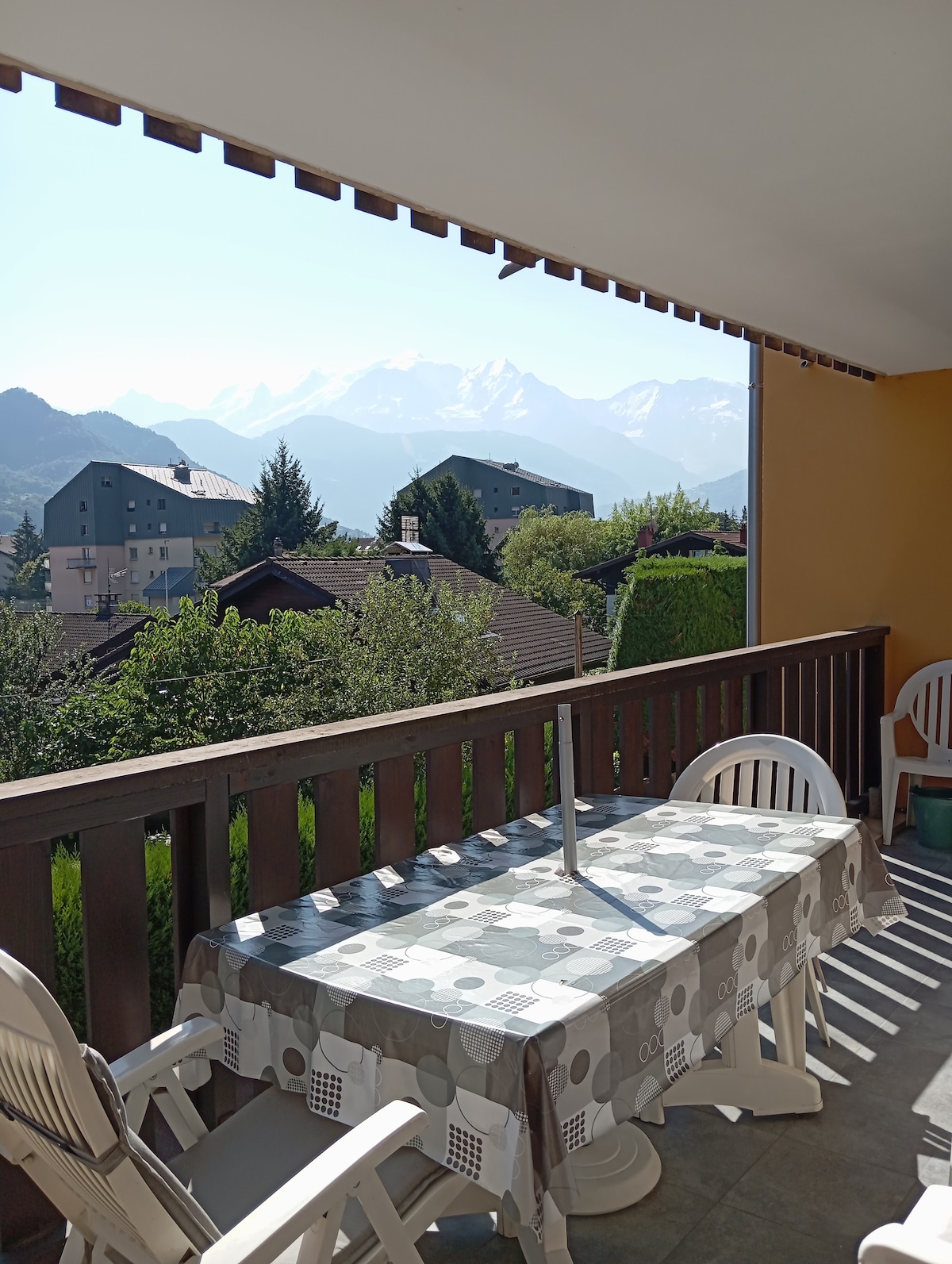 A balcony with a table set for outdoor dining is featured, accompanied by several white chairs. A scenic view of mountains and lush greenery is visible in the background, creating a serene outdoor atmosphere.