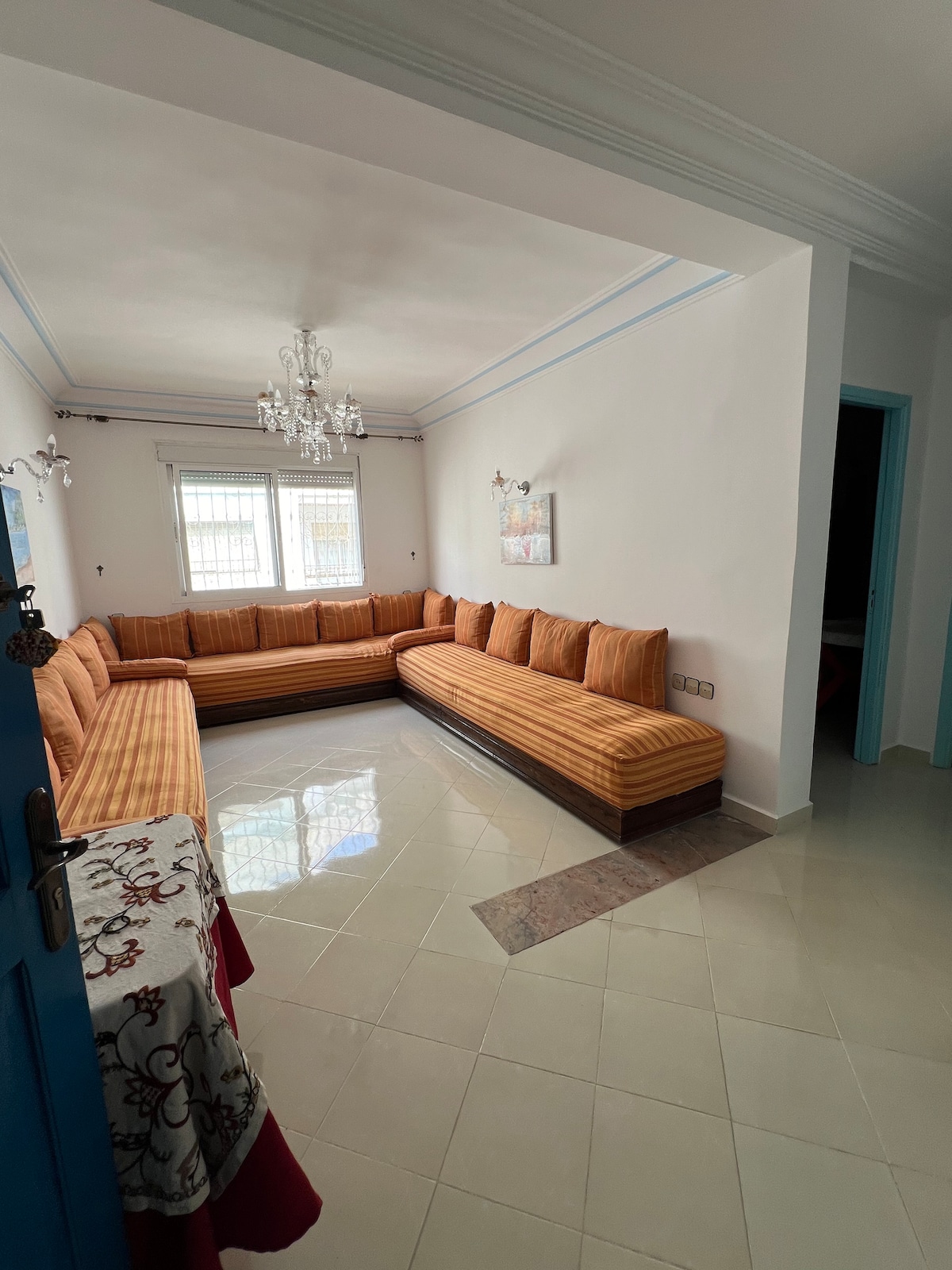 A spacious living area features a large, L-shaped sofa adorned with orange and cream stripes. Natural light fills the room through two windows, while a decorative chandelier hangs from the ceiling, adding a touch of elegance. The floor is tiled in light colors, enhancing the brightness.