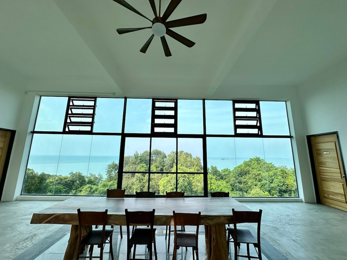 A spacious dining area is highlighted by a large wooden table surrounded by several chairs. Floor-to-ceiling windows offer an unobstructed sea view, while green trees and the horizon can be seen in the distance. A ceiling fan adds to the airy ambiance.