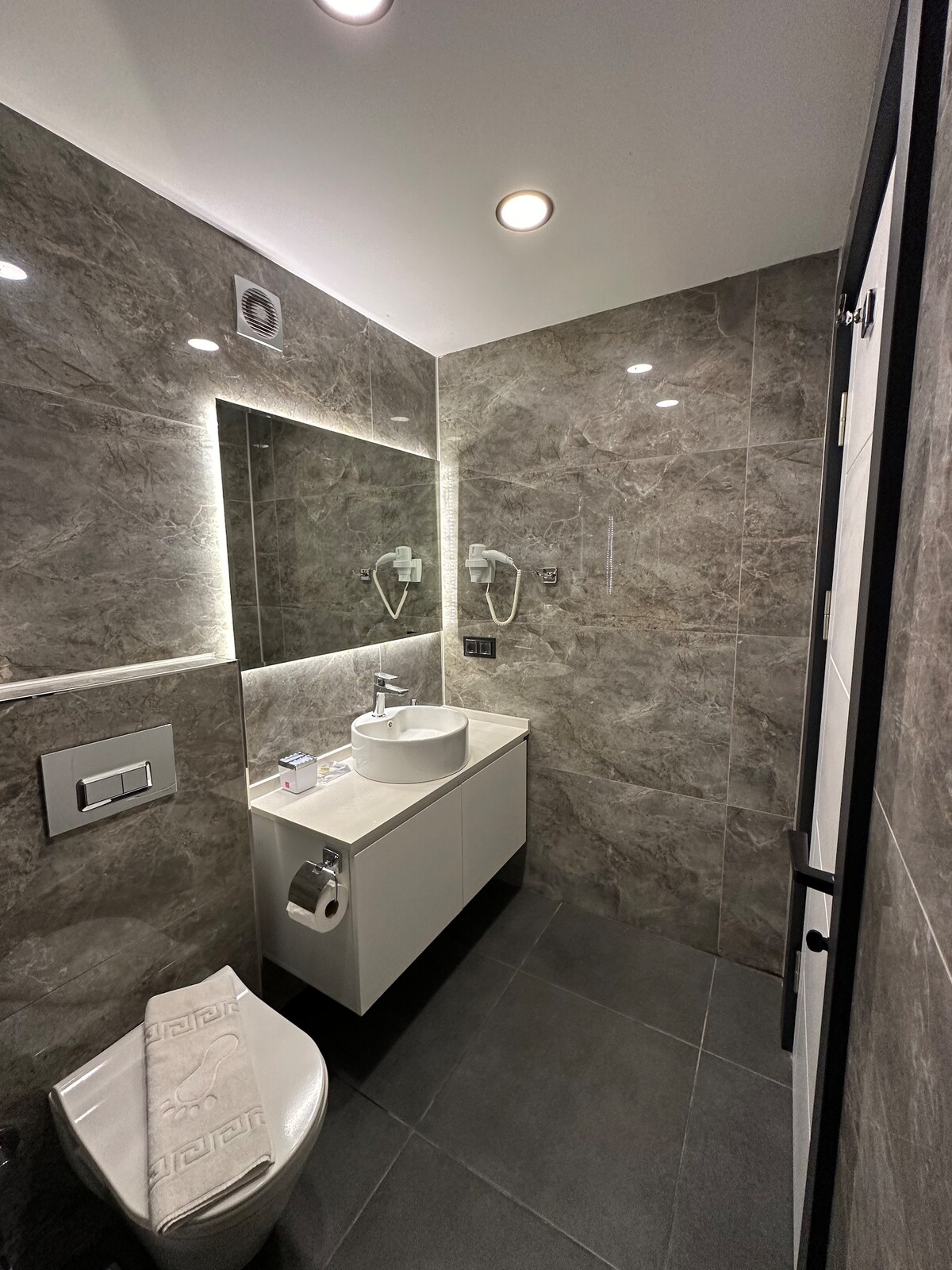 A modern bathroom is depicted, featuring sleek gray marble walls and a polished gray tiled floor. A sink with a minimalist design is positioned above a white vanity unit, with a large mirror illuminated by integrated lighting. Towel racks and toiletries are neatly arranged.