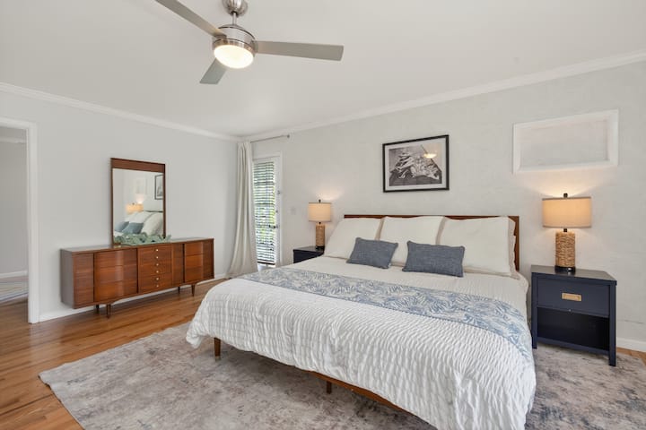 Bright and inviting bedroom featuring a plush king-size bed, soft lighting, hardwood floors, cozy seating area, mounted TV, and stylish coastal-inspired décor designed for comfort and relaxation.