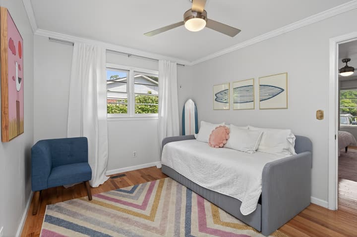 Bright coastal-inspired bedroom with a cozy daybed, crisp white linens, hardwood floors, and large windows. Beach-themed décor, colorful accents, and a comfortable chair create a relaxing, light-filled retreat.