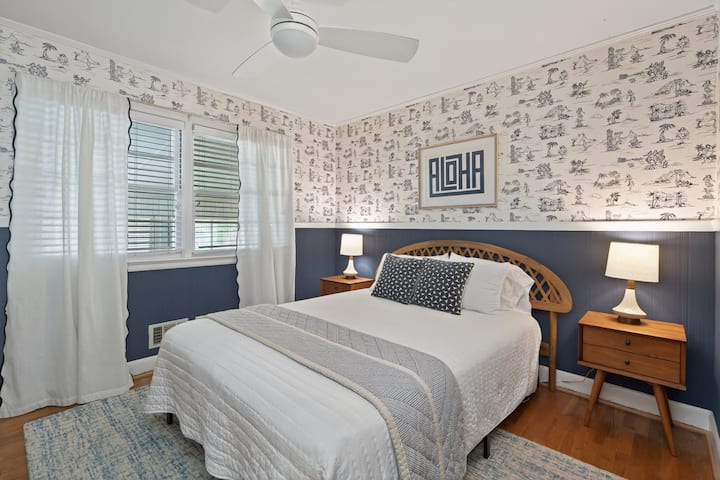 Coastal-inspired bedroom featuring a plush bed with crisp white linens, matching nightstands, warm wood accents, and an eye-catching ALOHA artwork set against charming tropical wallpaper.