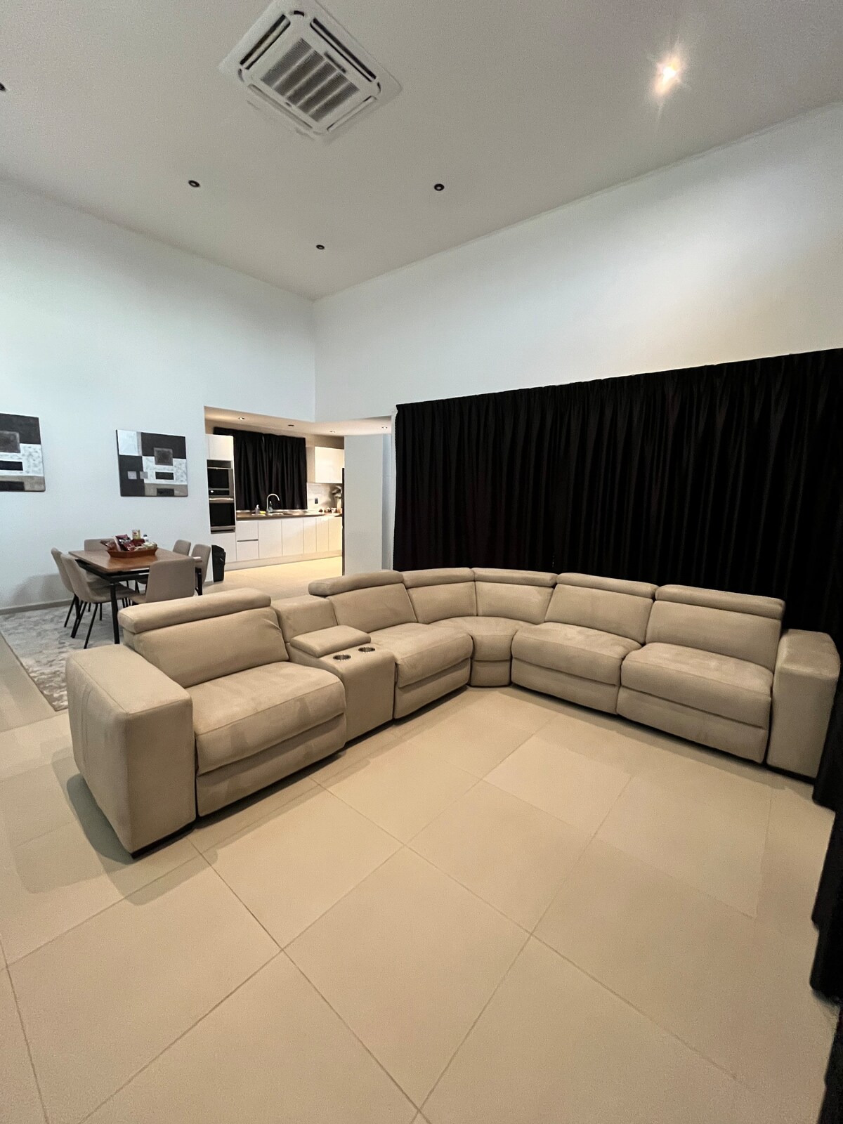 A spacious living area features a light-colored sectional sofa arranged in an inviting L-shape. Neutral flooring complements the contemporary design. The background reveals a dining area with a table and chairs, while sliding black curtains frame an entrance to the kitchen area.