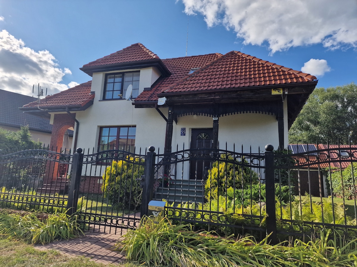 A charming house features a brick and stucco exterior with a distinctive red tiled roof. The front yard is enclosed by a decorative iron fence, showcasing well-maintained greenery and a walkway leading to the entrance. Bright skies and fluffy clouds complement the setting.