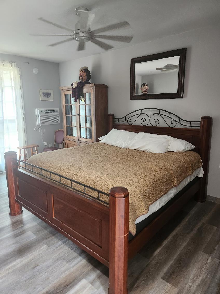 A spacious bedroom features a wooden bed with a decorative headboard, covered with a textured blanket. A large mirror hangs above the bed, reflecting soft light from nearby windows. A wooden display cabinet sits against the wall, and a ceiling fan provides air circulation.