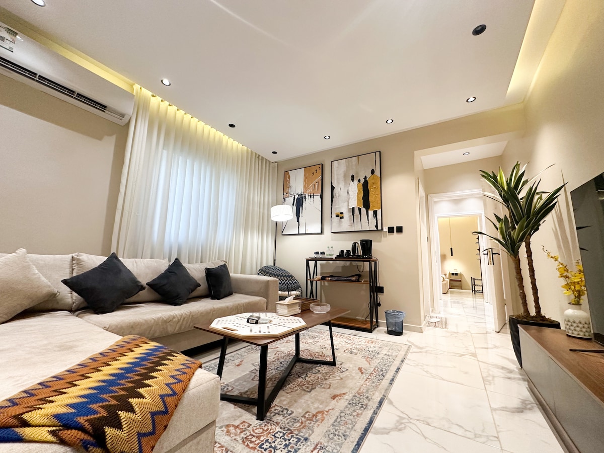 A modern living room features a beige sectional sofa adorned with black cushions. A coffee table with a decorative tray sits in front. Artwork is mounted on the wall, and a stylish lamp adds ambient lighting. Natural light filters through sheer curtains, enhancing the space.