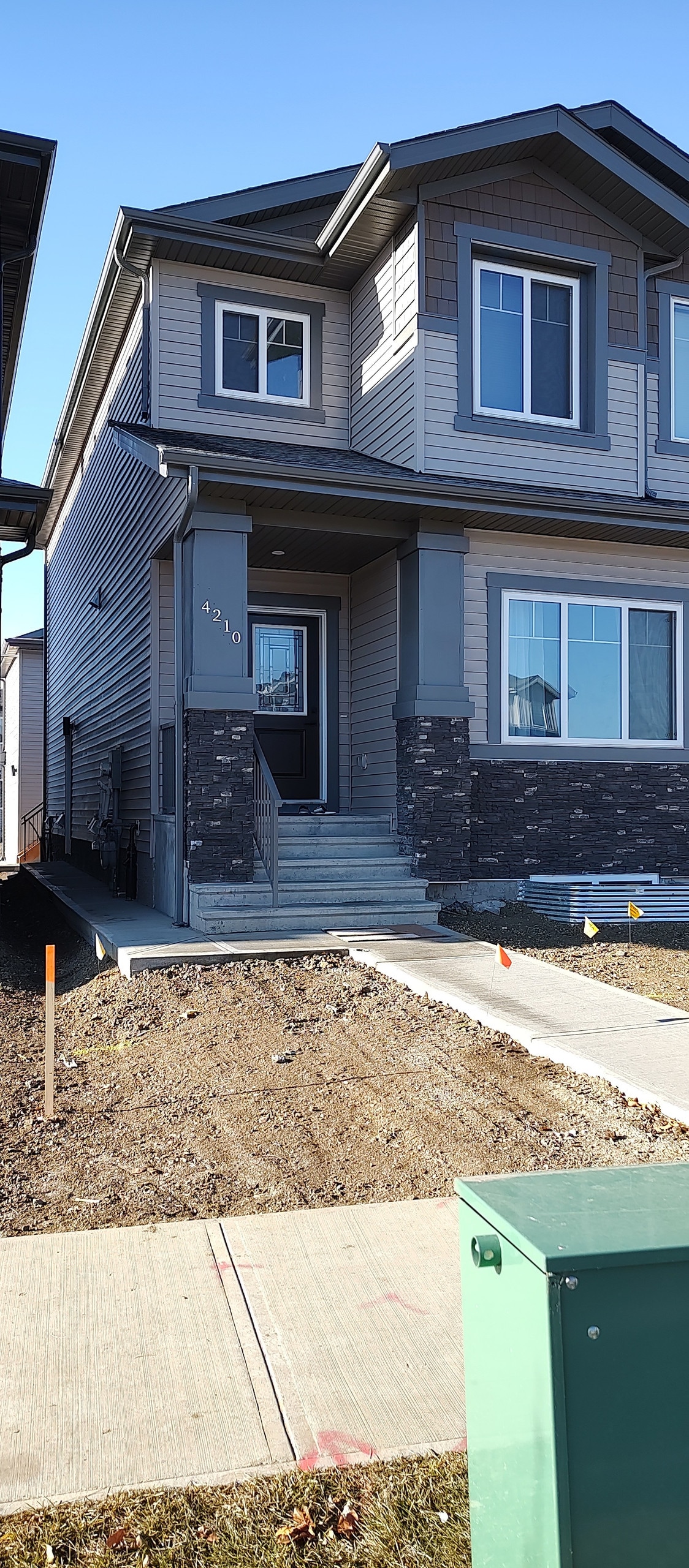 The exterior of a newly constructed home is depicted, featuring a modern facade with a mixture of siding and stone accents. The entrance includes a small set of steps leading to the front door, flanked by large windows that allow natural light to enter.