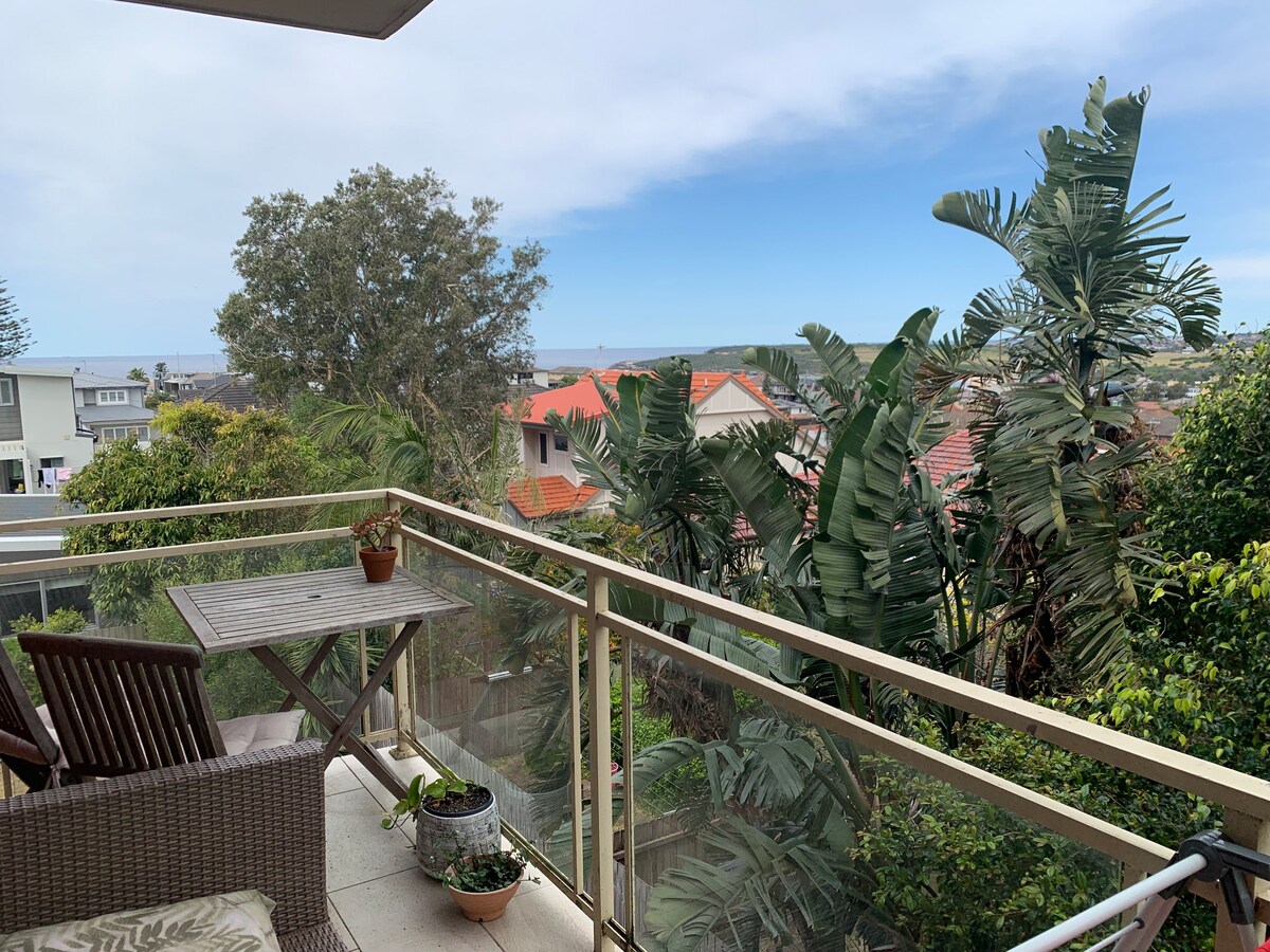 A private balcony is displayed, featuring a wooden table with two chairs and a potted plant. Lush greenery surrounds the area, while distant ocean views and palm trees are visible against a clear sky.