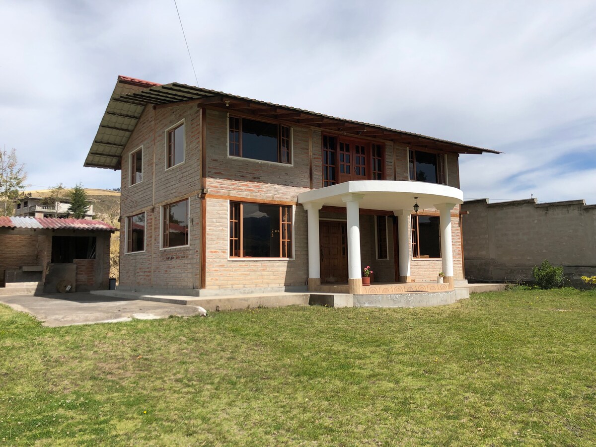 A two-story house with a blend of brick and wood design is displayed, featuring large windows for ample light. The entry is accentuated by a white columned porch, with a well-maintained grassy area surrounding the home, set against a clear sky.