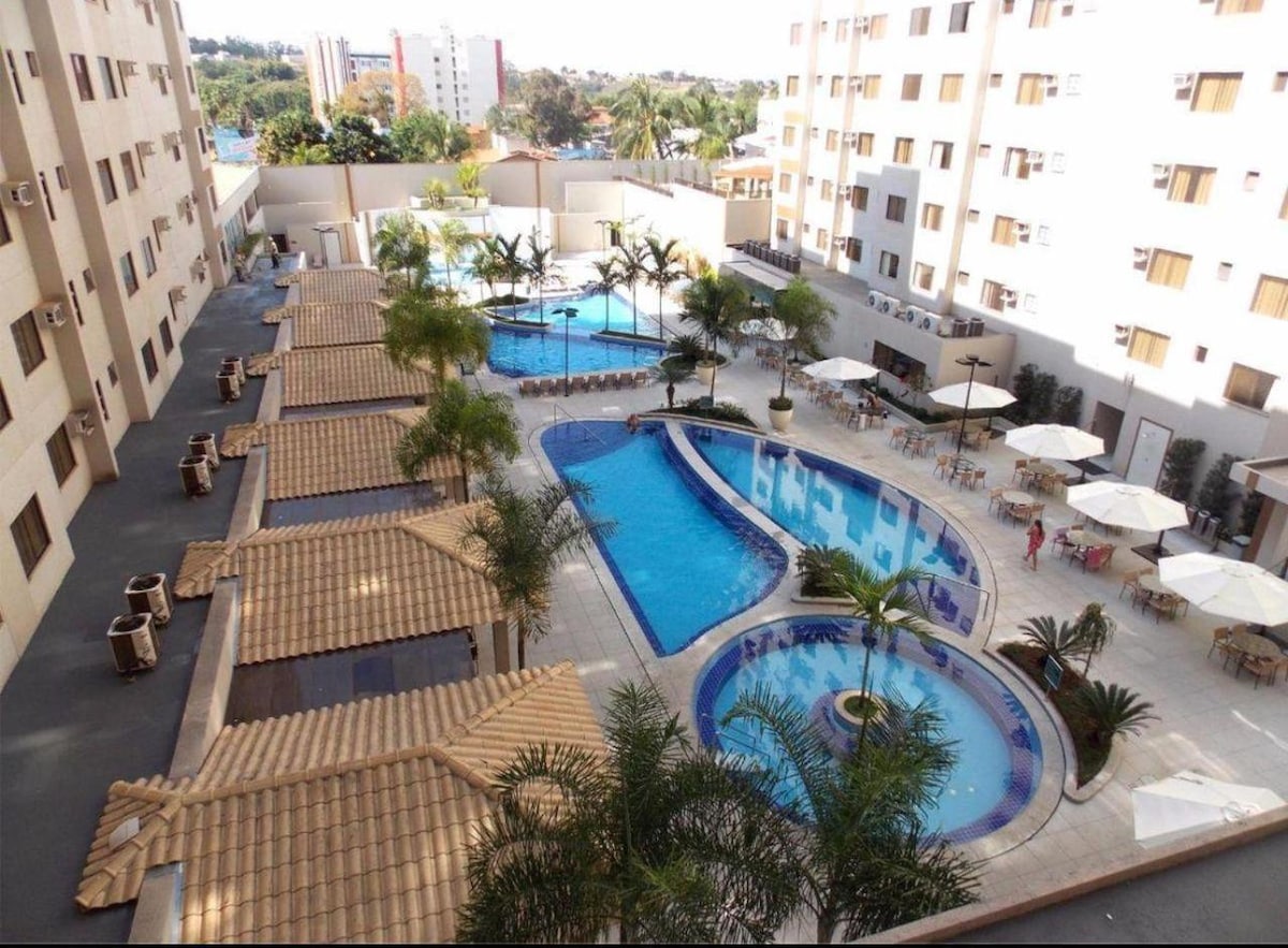 An aerial view showcases a spacious pool area surrounded by neatly arranged lounge chairs and umbrellas. Several palm trees provide greenery, and two swimming pools are visible, one featuring a circular design. The clean, modern architecture of the surrounding buildings complements the relaxing environment.