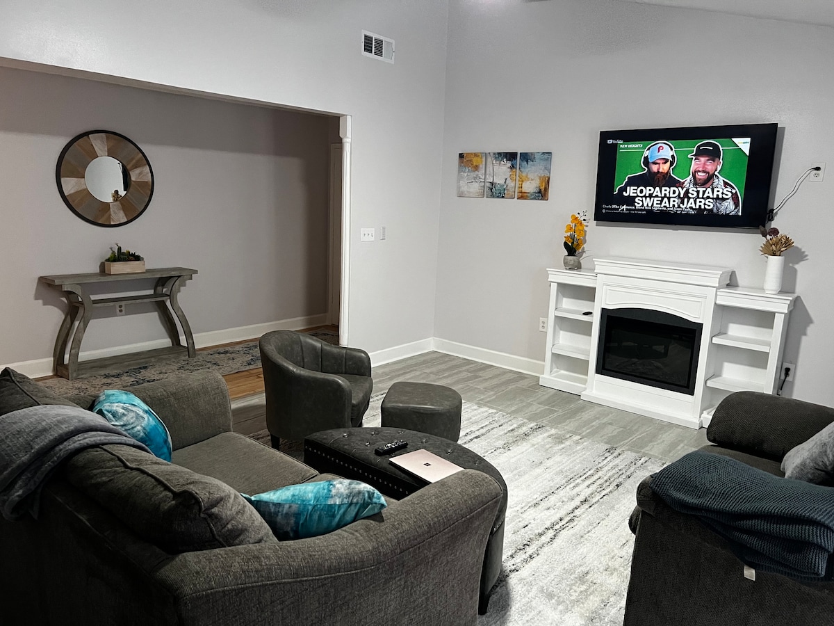 A stylish living area is furnished with a gray sectional sofa and a woven rug, enhancing the space's comfort. A fireplace is situated beneath a wall-mounted television, which displays a game show. Decorative elements include a round mirror and artwork on the walls.