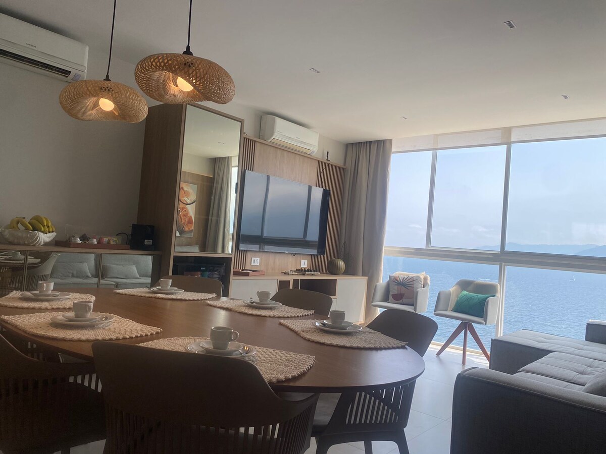 An open-concept living and dining area features a round wooden table surrounded by modern chairs. A large television is mounted on the wall opposite a panoramic window, showcasing a view of the sea. Soft pendant lights hang above the table, enhancing the tranquil atmosphere.