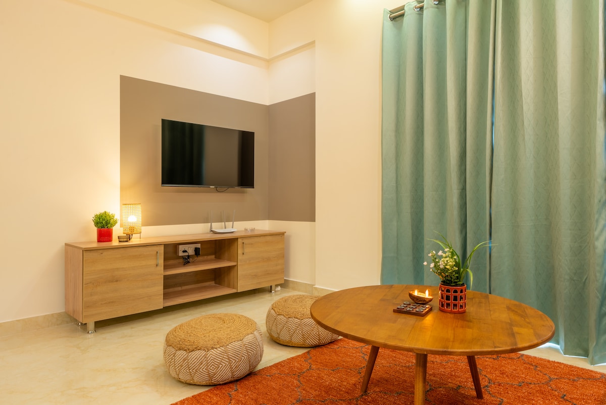A living area is presented with soft lighting and a flat-screen TV mounted on the wall. A circular wooden coffee table is centered on a textured orange rug, surrounded by round cushioned seating. Long curtains in a calming teal drape beside a decorative plant.