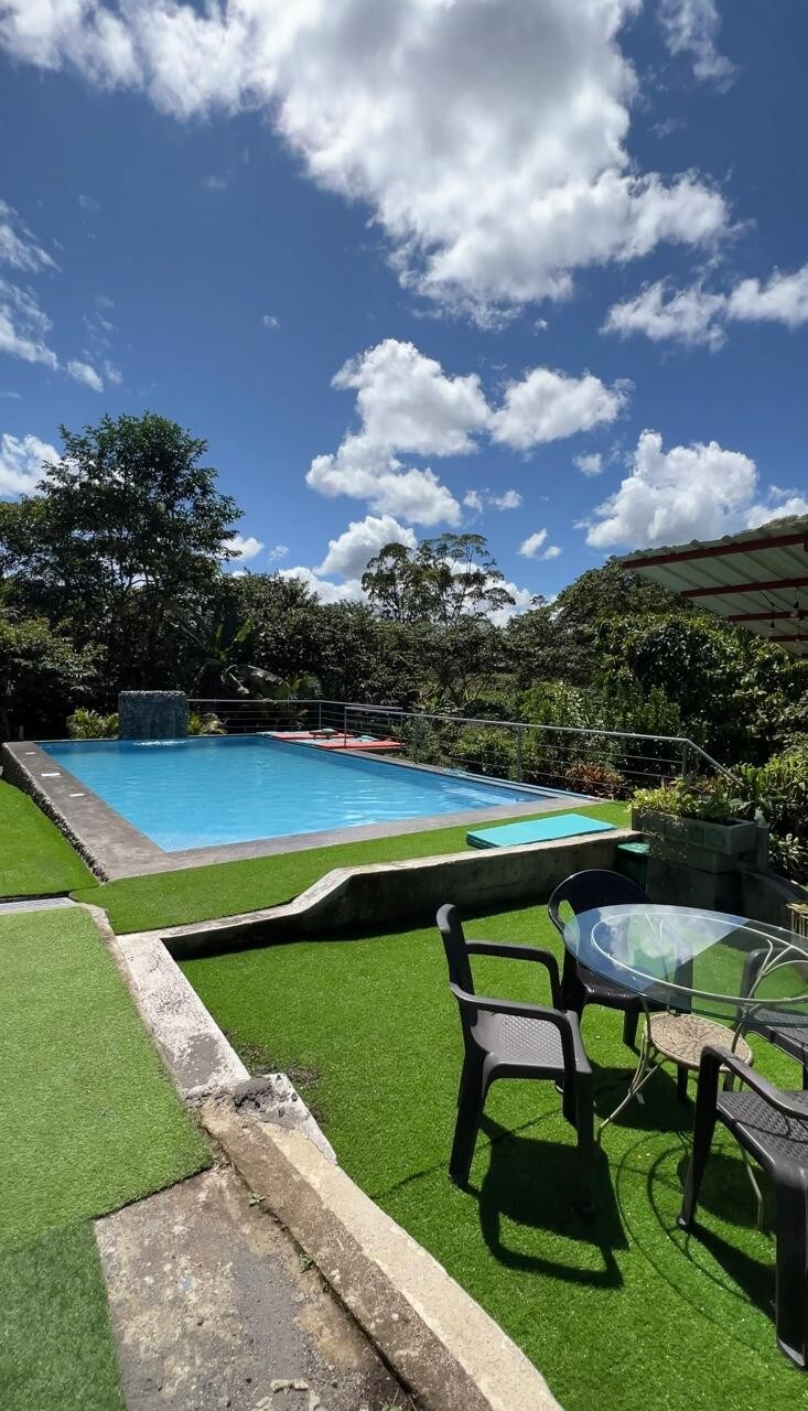 A serene outdoor scene features a large swimming pool surrounded by lush greenery. Sunlight illuminates the water, reflecting the bright blue sky filled with fluffy clouds. A table and chairs are positioned nearby on a grassy area, creating a comfortable spot for relaxation.
