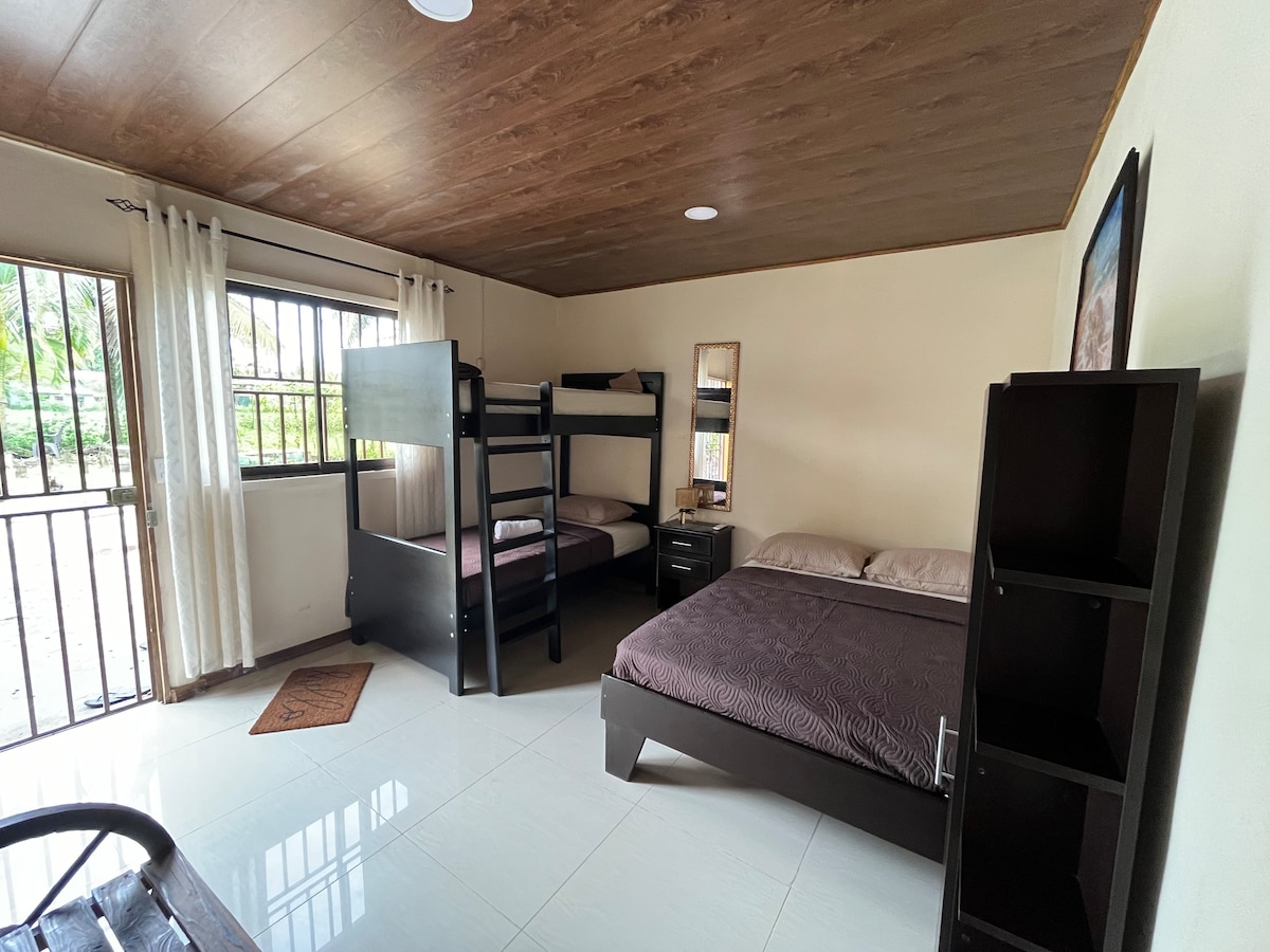 A new guest room is equipped with a spacious bed and a bunk bed, accommodating four guests. Natural light enters through the window, which is fitted with sheer curtains. A nightstand and a small cabinet are positioned beside the beds, enhancing functionality.