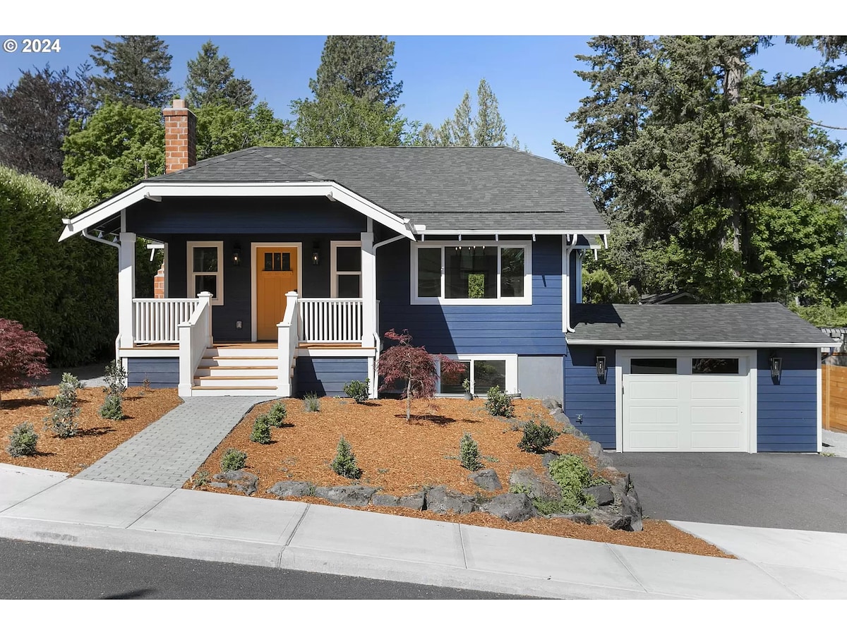 A charming two-story home features a blue exterior with white trim and a welcoming front porch. Lush landscaping surrounds the entrance, with neatly arranged stones and colorful plants enhancing the curb appeal. A single-car garage is visible on the right side of the image.