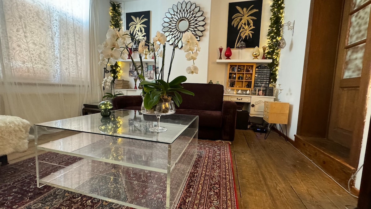 The living area features a deep brown sofa positioned beside a clear, glass coffee table. Elegant white orchids are arranged in a vase atop the table. The space is adorned with wall art depicting palm trees, alongside decorative lighting and a textured rug on the hardwood floor.