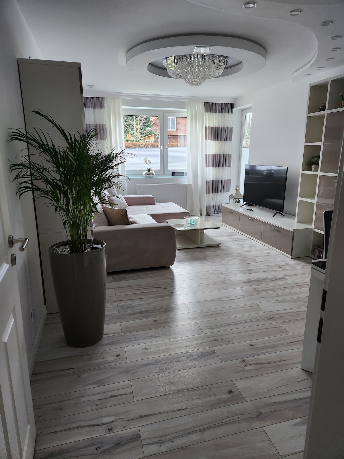A bright living area features a sleek chandelier and large windows that allow natural light to enter. A comfortable couch is positioned opposite a television on a light-colored media unit. Decorative plants are placed in a tall pot, adding greenery to the space.