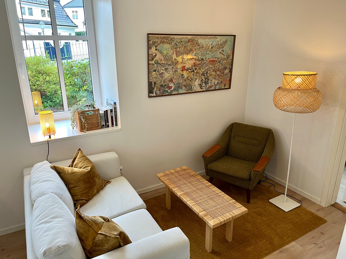 A bright living area is presented, featuring a white sofa adorned with gold cushions. A natural wood coffee table sits on a woven rug, complemented by a green accent chair. Soft light filters through a nearby window, highlighting a colorful piece of art on the wall.
