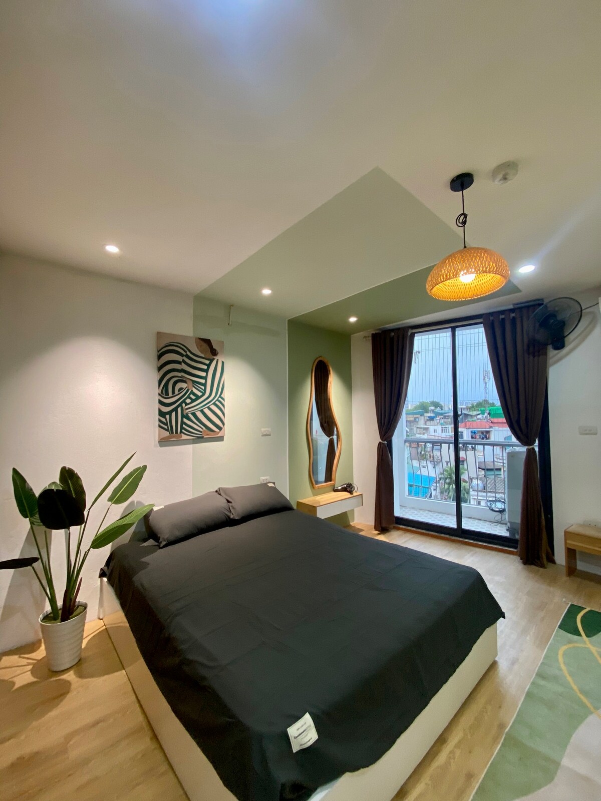 A modern bedroom features a spacious bed covered with dark linens, complemented by a stylish light fixture. Natural light enters through a large glass door leading to a balcony. A decorative mirror and a green plant enhance the contemporary aesthetic.