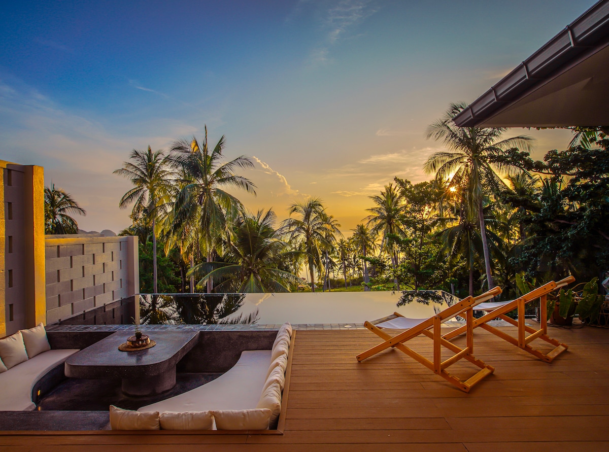 A spacious terrace features comfortable loungers positioned to enjoy sunset views. Lush palm trees and ocean vistas create a tranquil backdrop, while a modern seating area invites relaxation. The warm tones of the wood deck enhance the serene atmosphere.