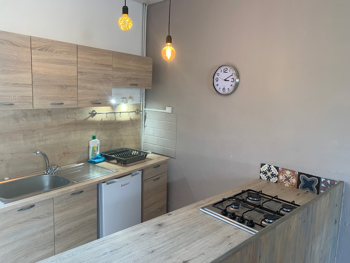 The kitchen features modern wooden cabinetry and a gas stove with five burners. A stainless steel sink is positioned under wall-mounted lighting, while a clock is displayed on the wall beside the fridge. Decorative tile elements add color to the kitchen's design.