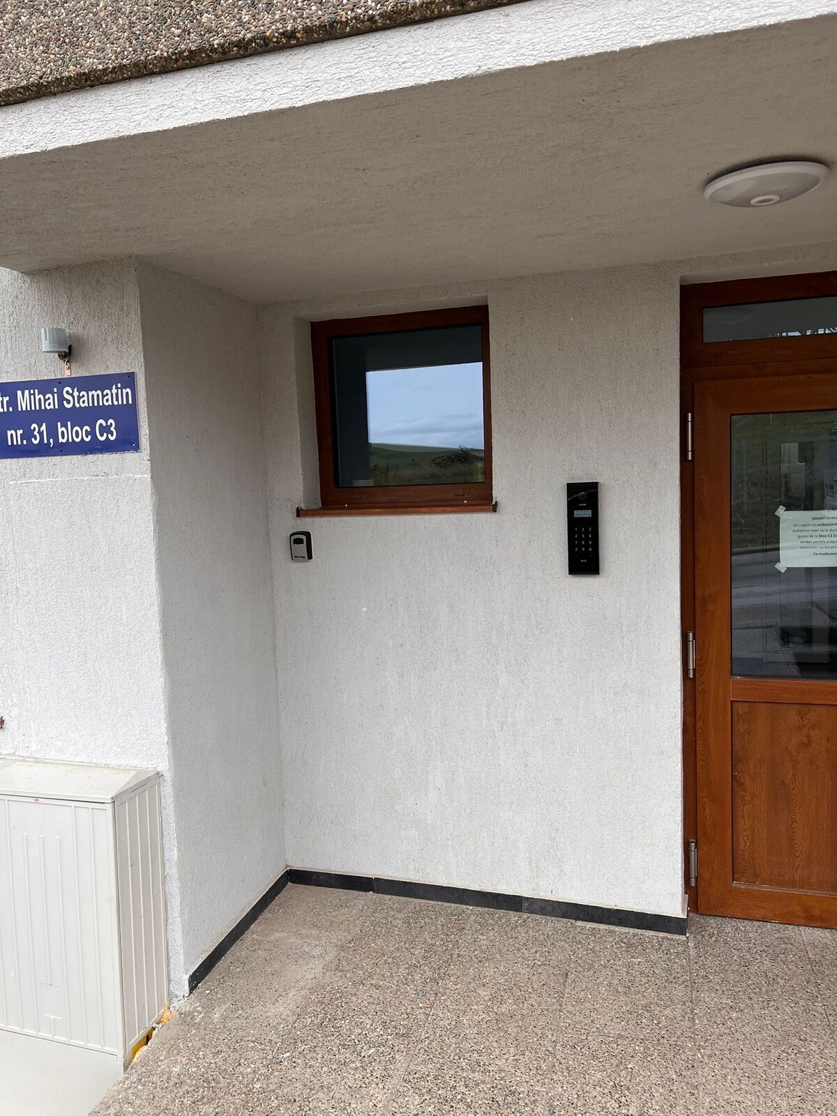 A modern entrance is shown, featuring a solid wooden door with a sleek digital intercom panel. The white textured walls provide a neutral backdrop, and a small window above the door allows natural light to enter. A sign displaying the address is mounted beside the entrance.