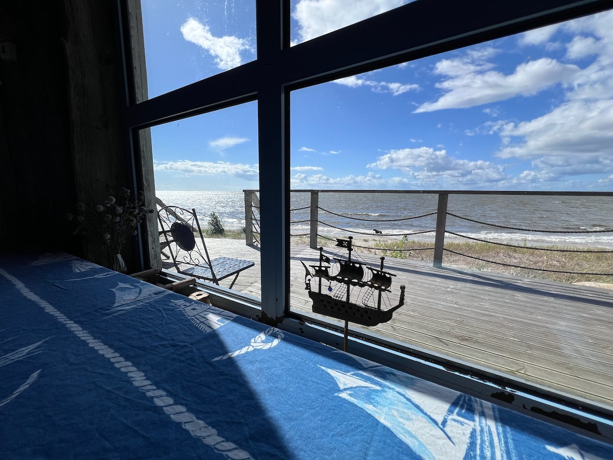 A serene view from a window captures shimmering water and a distant deck overlooking the beach. A decorative ship model sits on a table, with sunlight illuminating the scene and clouds drifting across a bright blue sky.