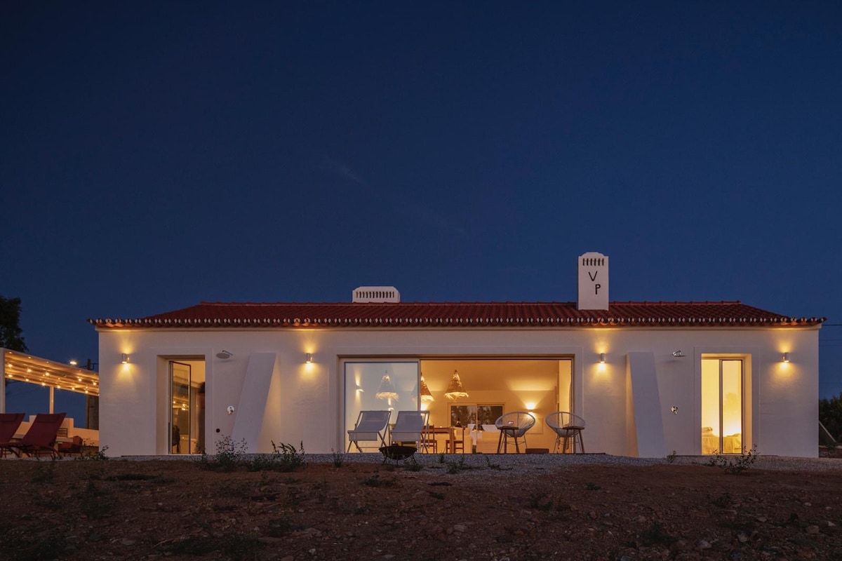 A spacious house is shown at dusk, featuring large glass doors that illuminate the interior. The exterior is framed by a red-tiled roof and soft outdoor lighting, enhancing the serene atmosphere of the natural landscape surrounding the property.