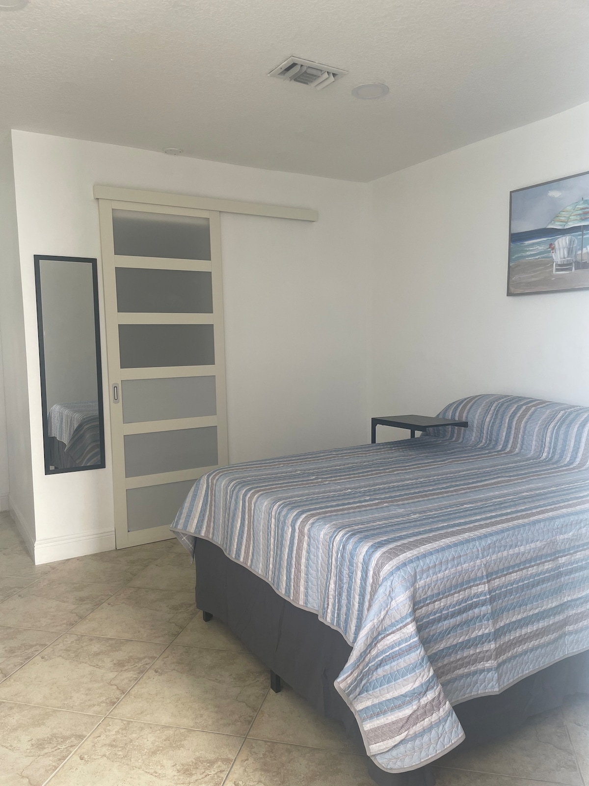 A bright sleeping area features a striped bedspread over a spacious bed. A bedside table stands next to the bed, while a full-length mirror and a glass-paneled door create a sense of openness. Light-colored walls and tiled flooring enhance the room's inviting ambiance.
