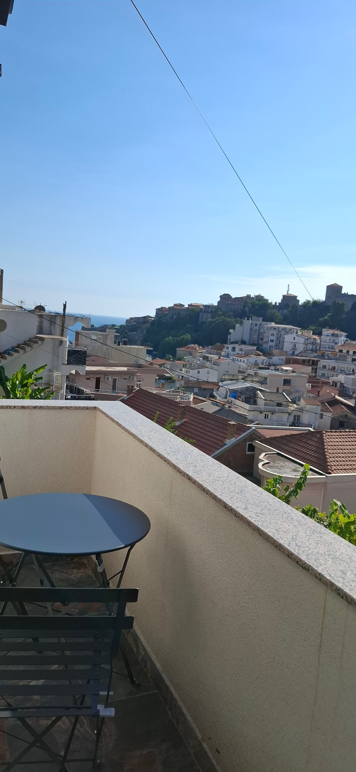 A small balcony is visible, featuring a round table and a pair of chairs. A panoramic view of a hillside town can be seen, with rooftops extending towards a clear blue sky. Lush greenery is present along the balcony's edge.
