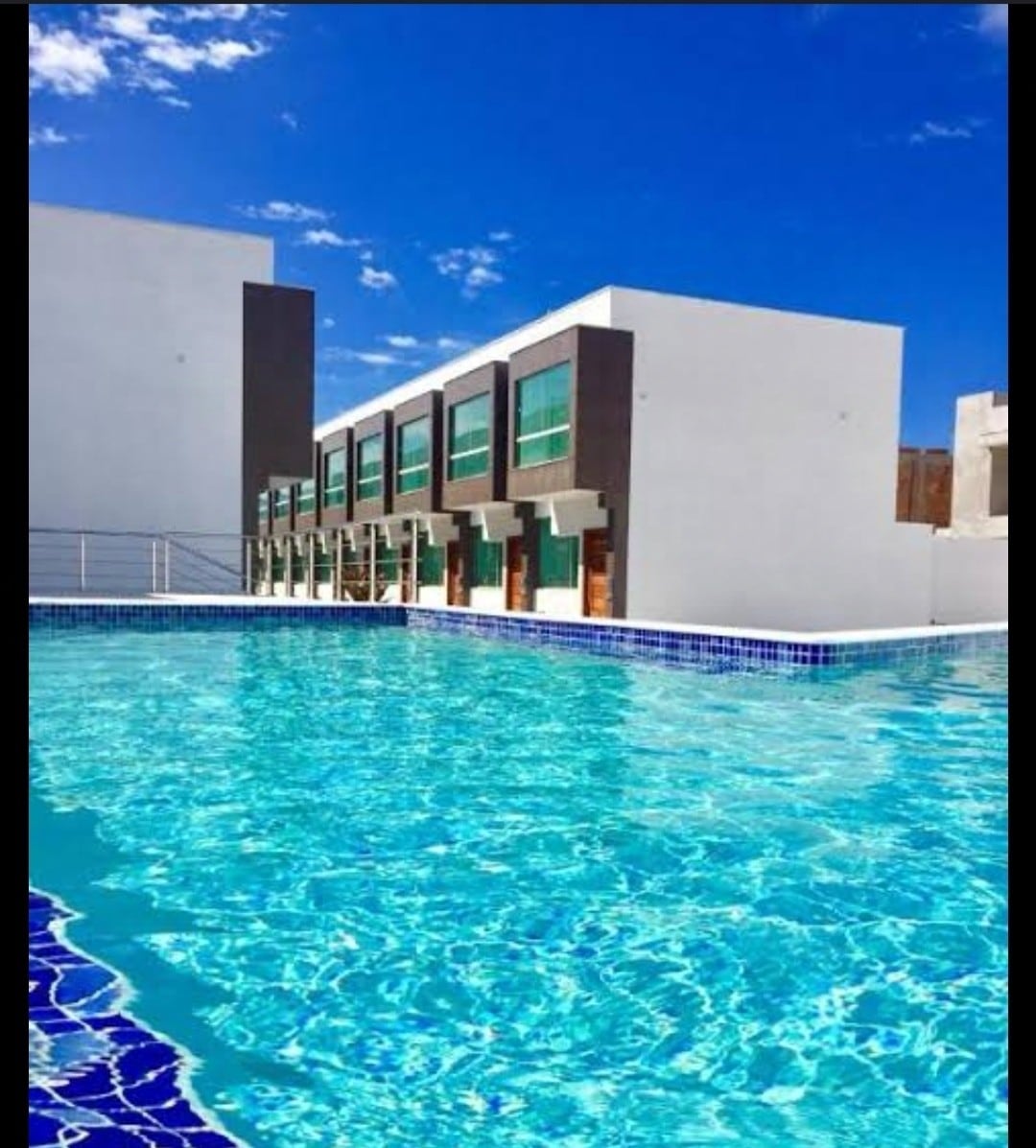 A clear and inviting swimming pool features shimmering blue water, reflecting the bright sky. The pool area is surrounded by modern, two-story buildings with large windows, providing a contemporary atmosphere for relaxation and leisure.