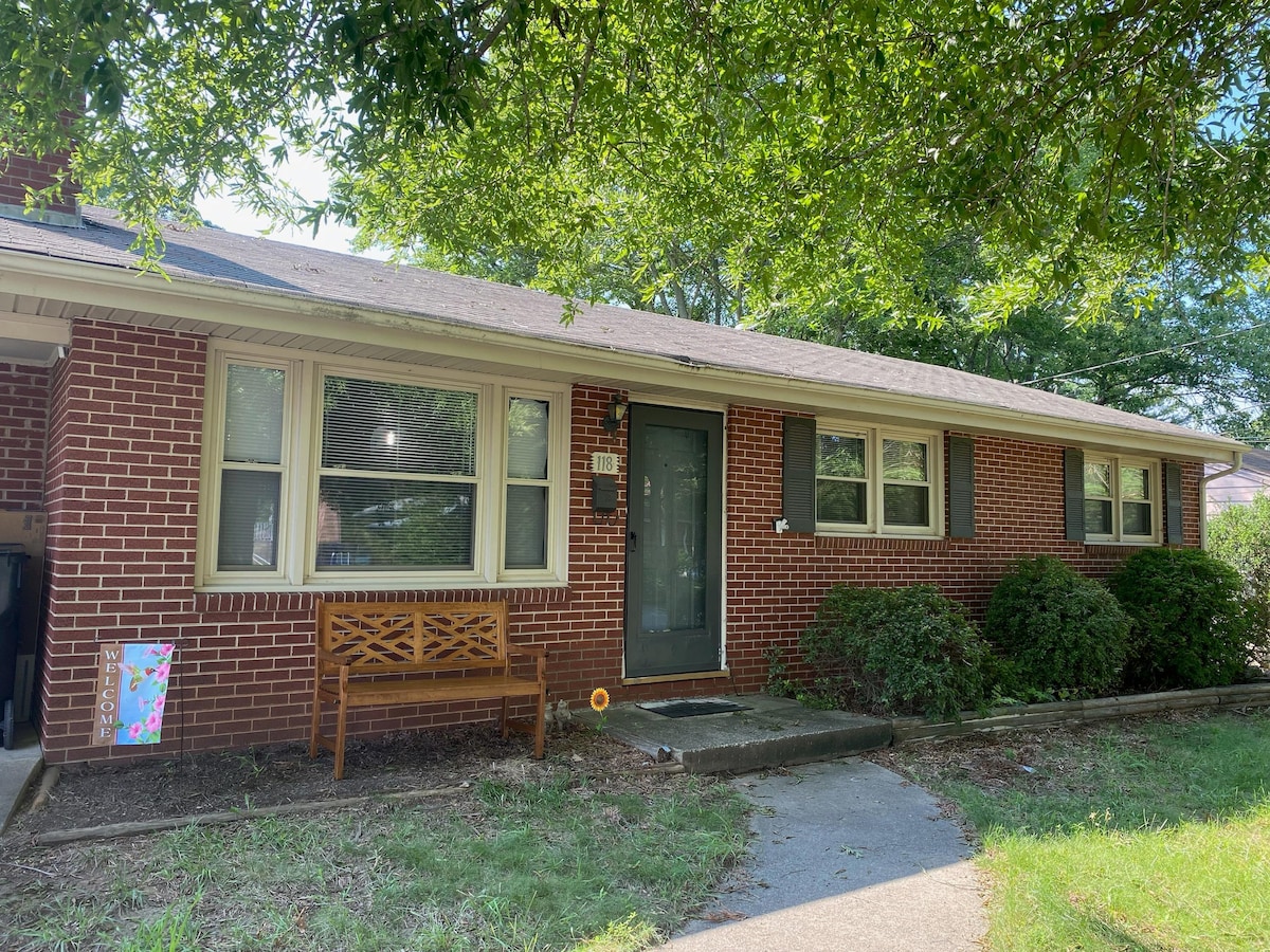 A single-story brick home is framed by lush greenery, featuring a welcoming front porch with a wooden bench. Large windows allow natural light to fill the space, while the level pathway leads to the front door. Neatly landscaped bushes enhance the inviting entrance.
