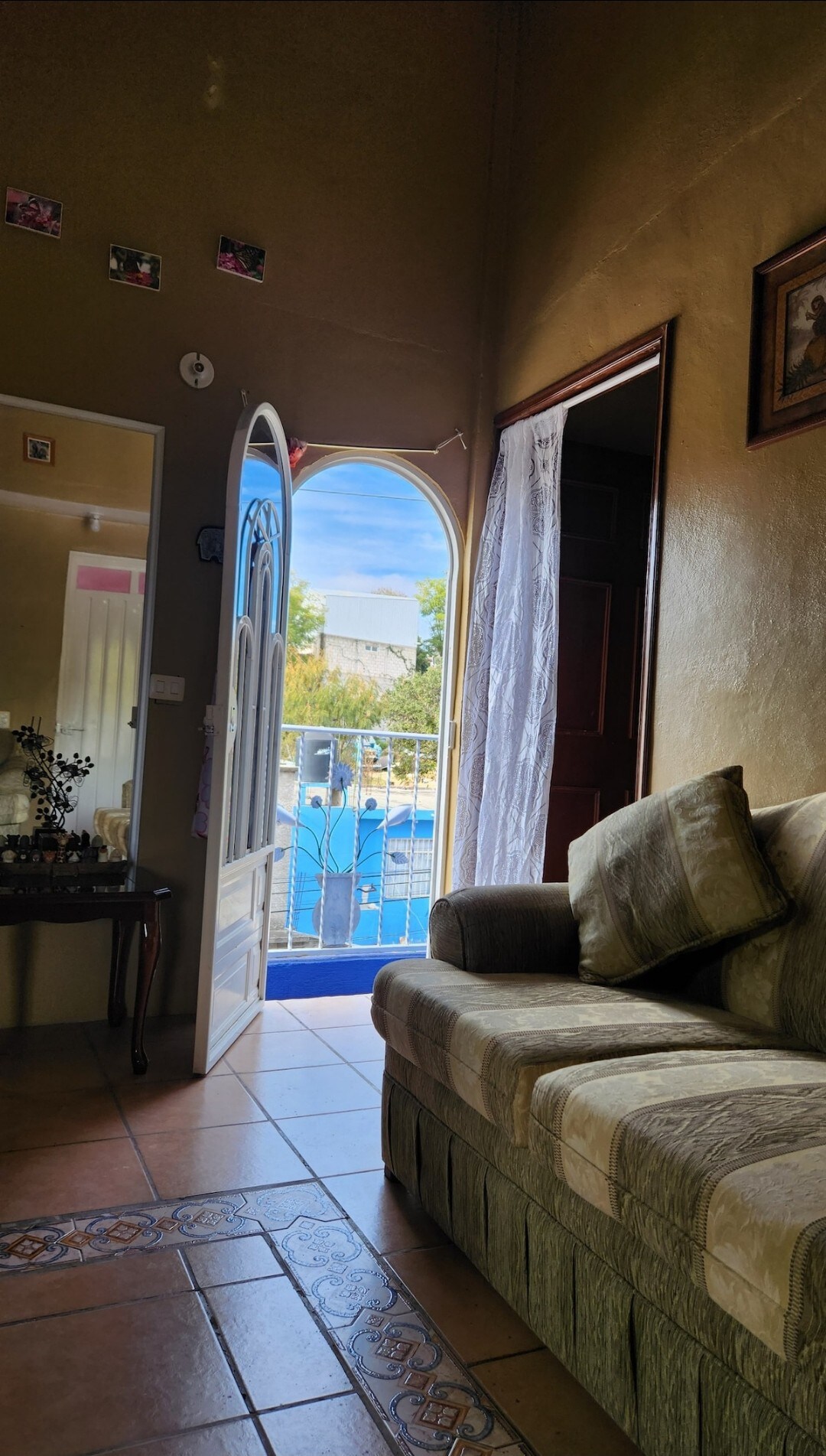 A cozy living area features a comfortable sofa positioned near an arched doorway. Sunlight filters through the open door, which leads to a view of a swimming pool outside. Soft tones and decorative pieces add character to the room.