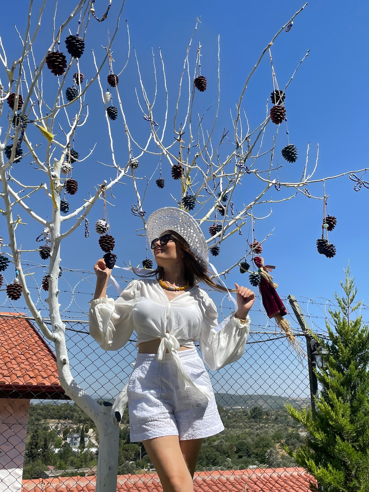 A white tree adorned with pine cones stands against a clear blue sky. A person poses beneath the tree, dressed in light-colored clothing and a wide-brimmed hat. In the background, a green landscape is visible along with a red-tiled roof.