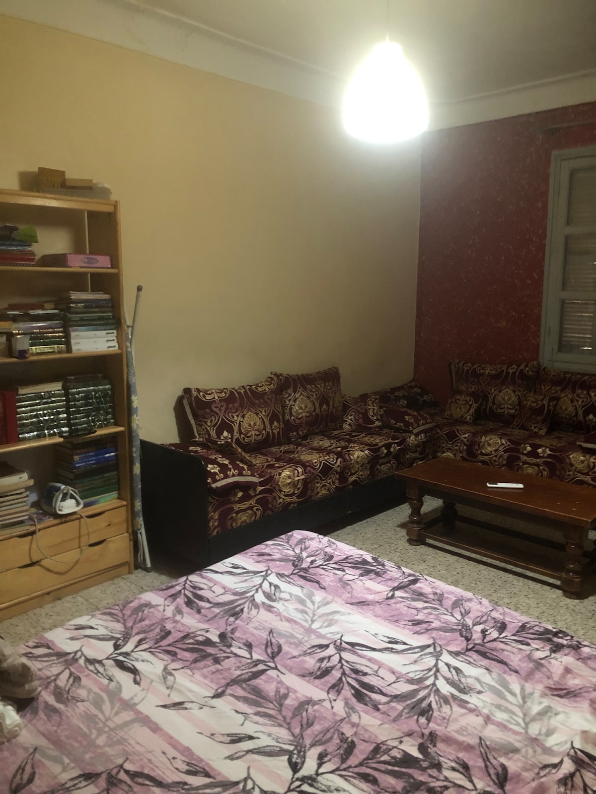 A cozy living area is furnished with patterned sofas and a wooden coffee table. A bookshelf filled with various volumes stands against one wall. Soft lighting is provided by a hanging lamp above the seating area, while a light-colored floral bedspread is visible in the foreground.