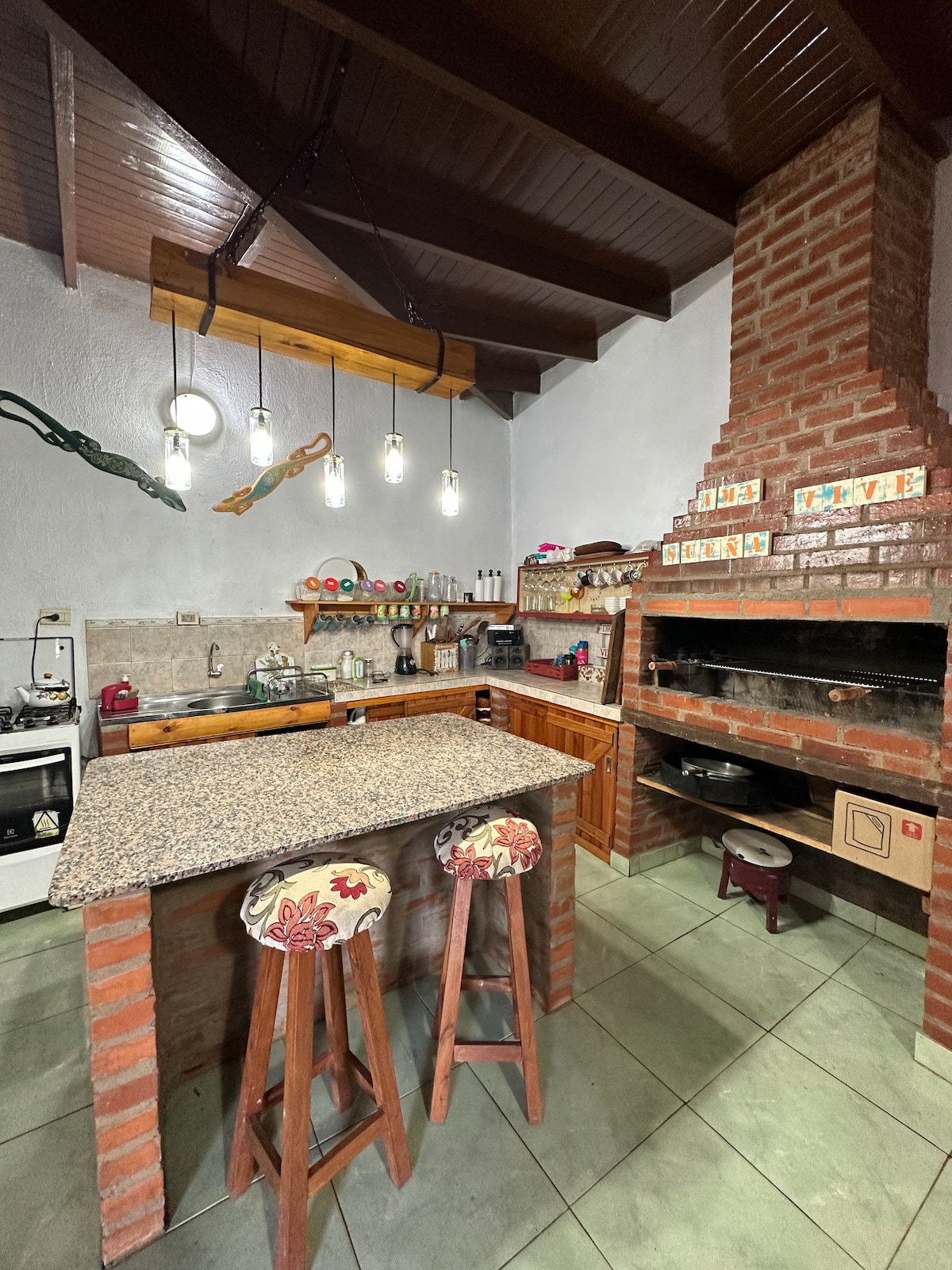 An inviting kitchen space features a granite countertop with two wooden stools. Open shelves display colorful dishware, while a brick barbecue area is positioned against the back wall. Unique light fixtures hang from the ceiling, enhancing the room's rustic charm.