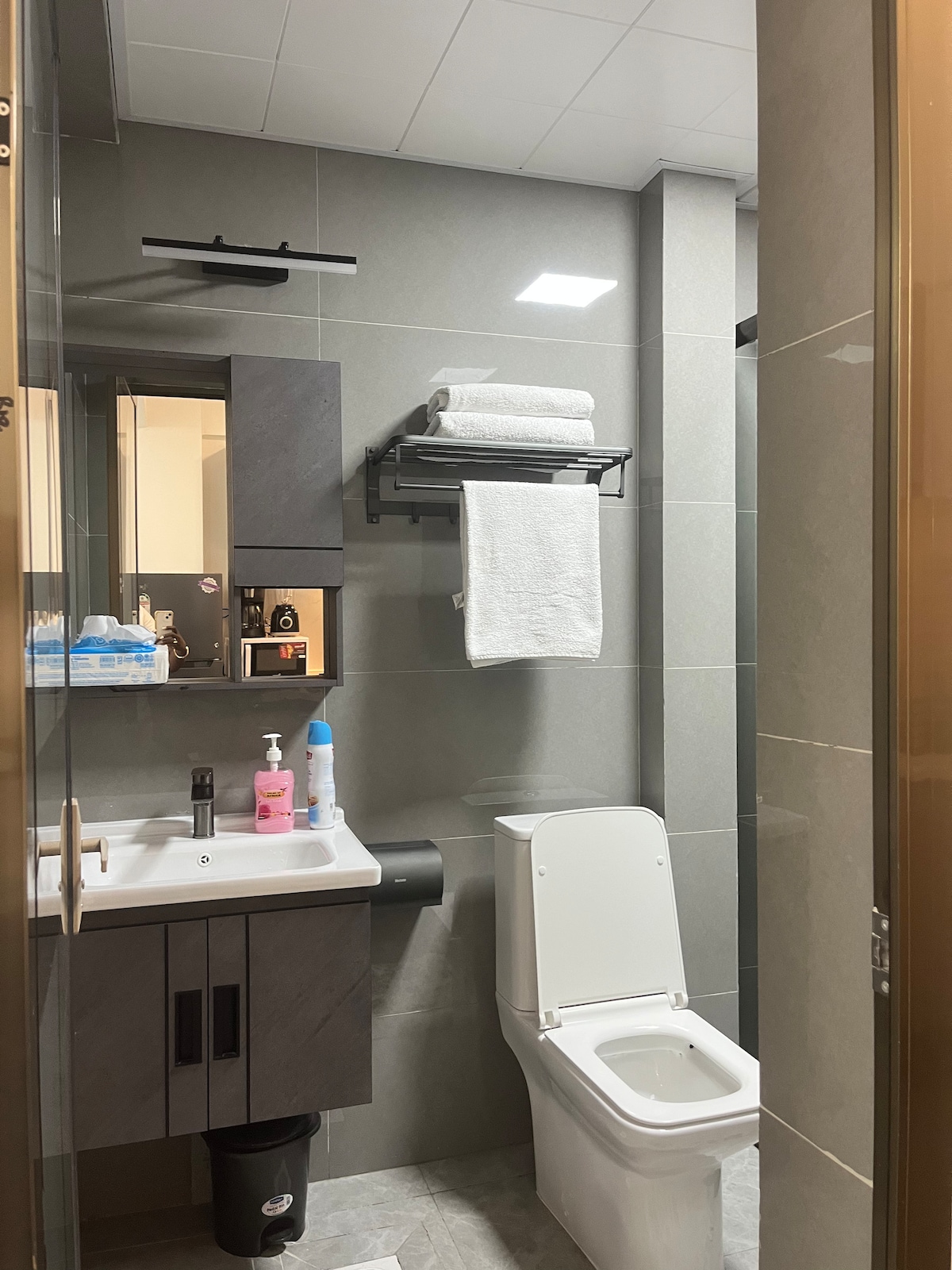 A modern bathroom features sleek gray tiles and minimalistic cabinetry. A sink with a mirror is positioned on the left, while a white toilet is visible on the right. Towels are neatly arranged on a wall-mounted rack above the sink, offering convenience and organization.