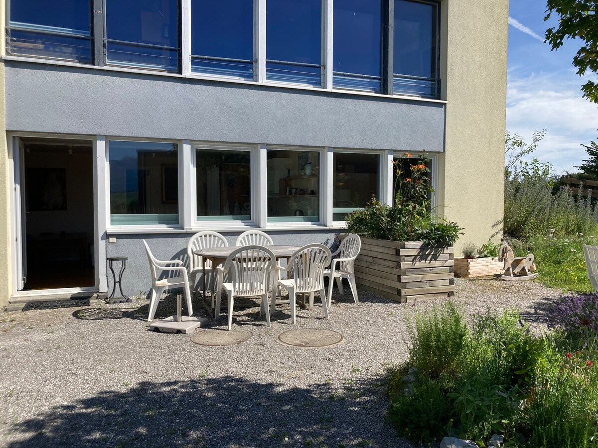 An outdoor patio area features a large table surrounded by several white chairs. Natural light fills the space from large windows, offering views of the garden. Stone pathways and planters with greenery define the area, creating a serene and inviting setting.