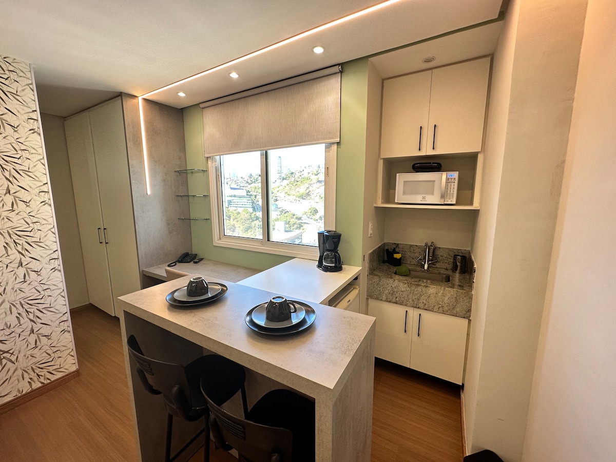 A modern kitchen is equipped with essential appliances, including a microwave and coffee maker. A stone countertop serves as a breakfast bar with two black stools positioned beneath it, while a window allows natural light to illuminate the space.