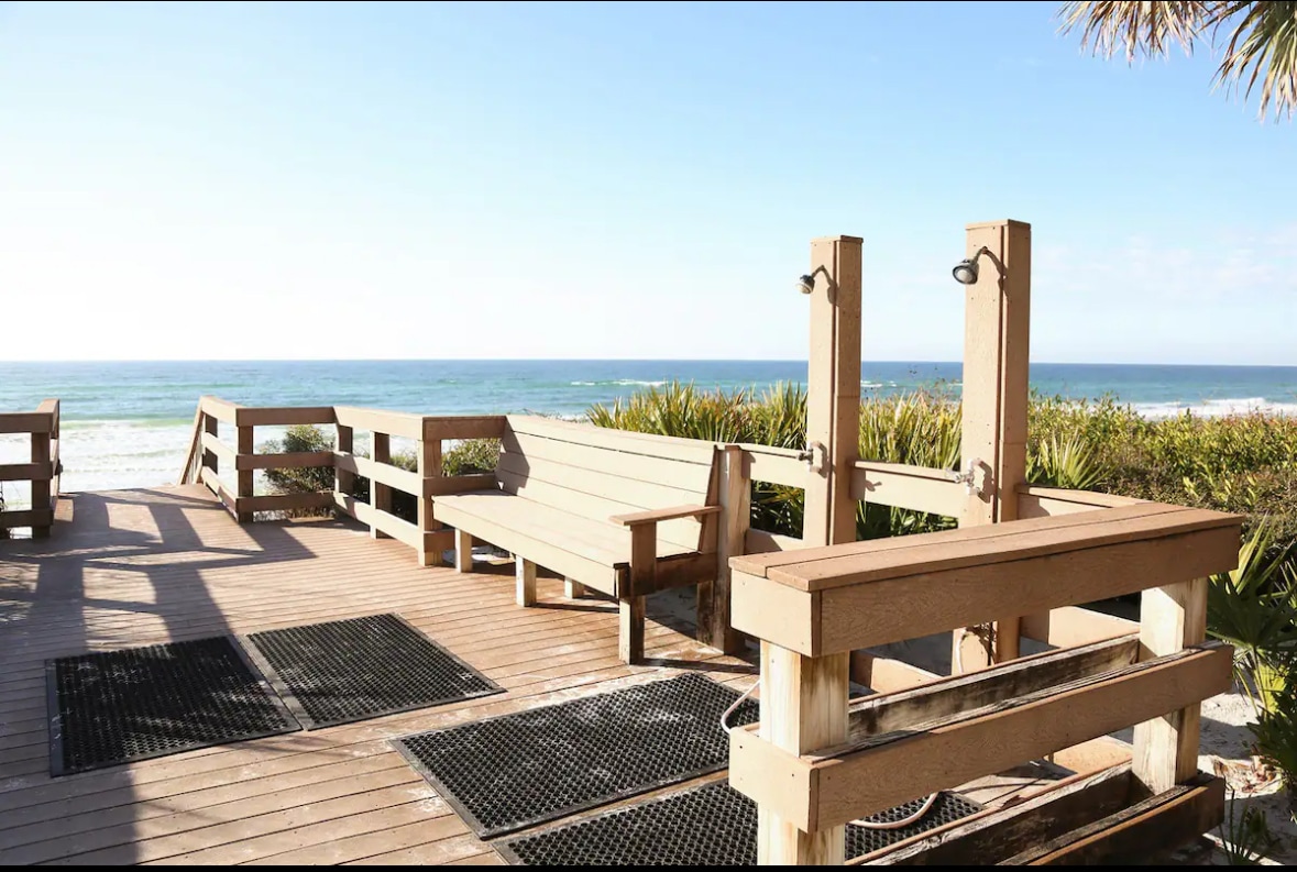 The image depicts a wooden deck leading to the beach, featuring benches and outdoor showers for convenience. A clear view of the ocean, where the water meets the horizon, is visible. Sunlight brightens the area, creating a welcoming outdoor space.